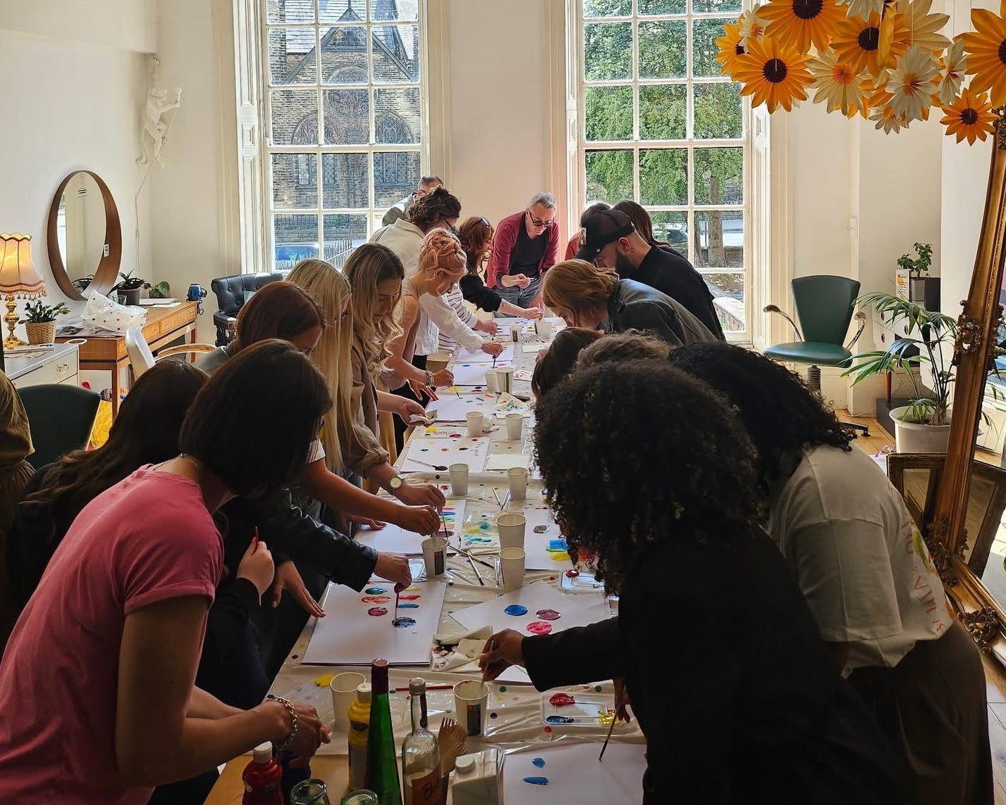 Group of people stood around a long table in a room with large windows and sunflower decorations painting onto sheets of paper.